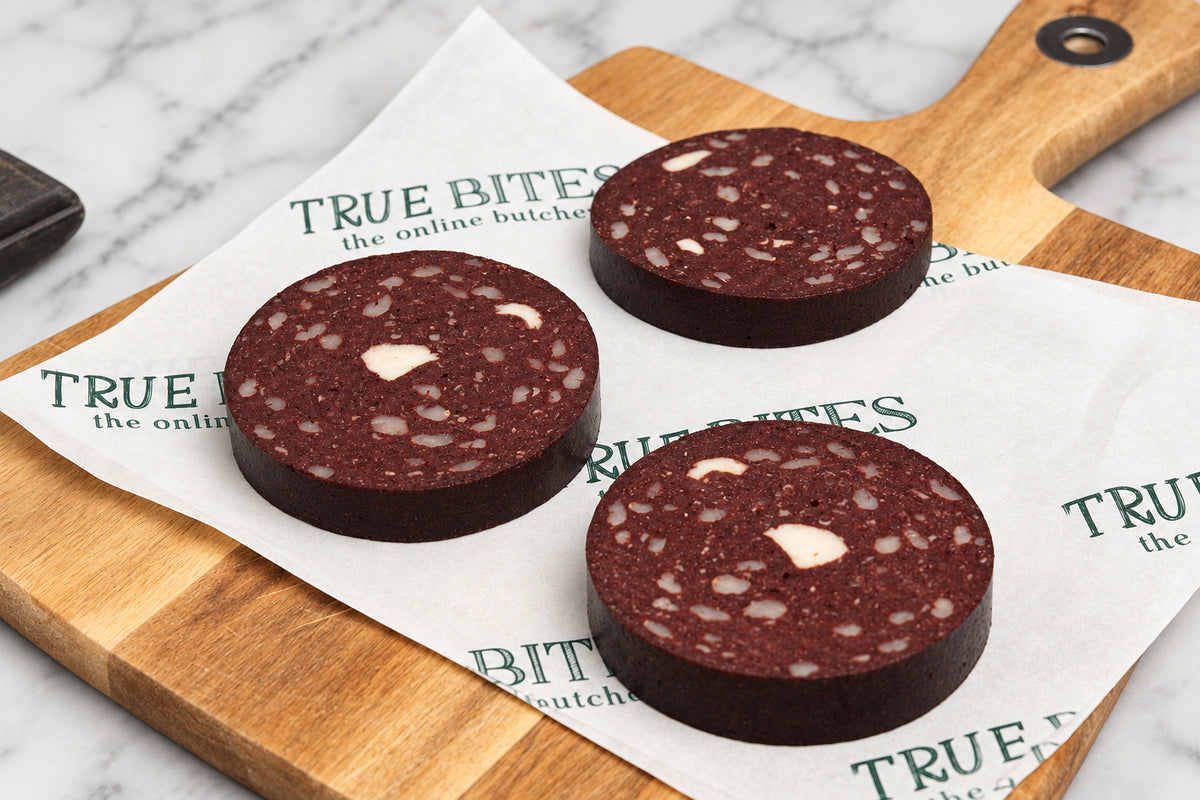 Three round slices of black pudding with white fat marbling, arranged on white paper printed with the brand name 'TRUE BITES' on a wooden cutting board.