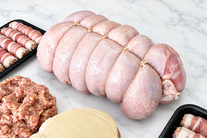 A tied raw turkey breast roast on a white cutting board, surrounded by packaged pigs in blankets, 450g of sausage meat.