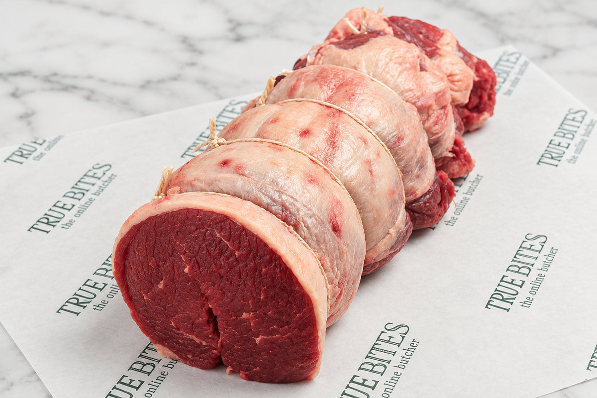 A raw rolled beef brisket tied with string and resting on branded paper, with a meat thermometer visible in the background on a dark wooden surface.