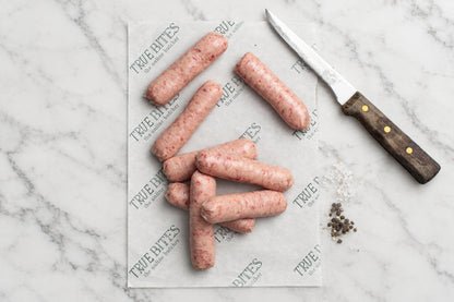 8 Raw pork sausages arranged on branded parchment paper with a knife and coarse salt on a dark wooden surface.