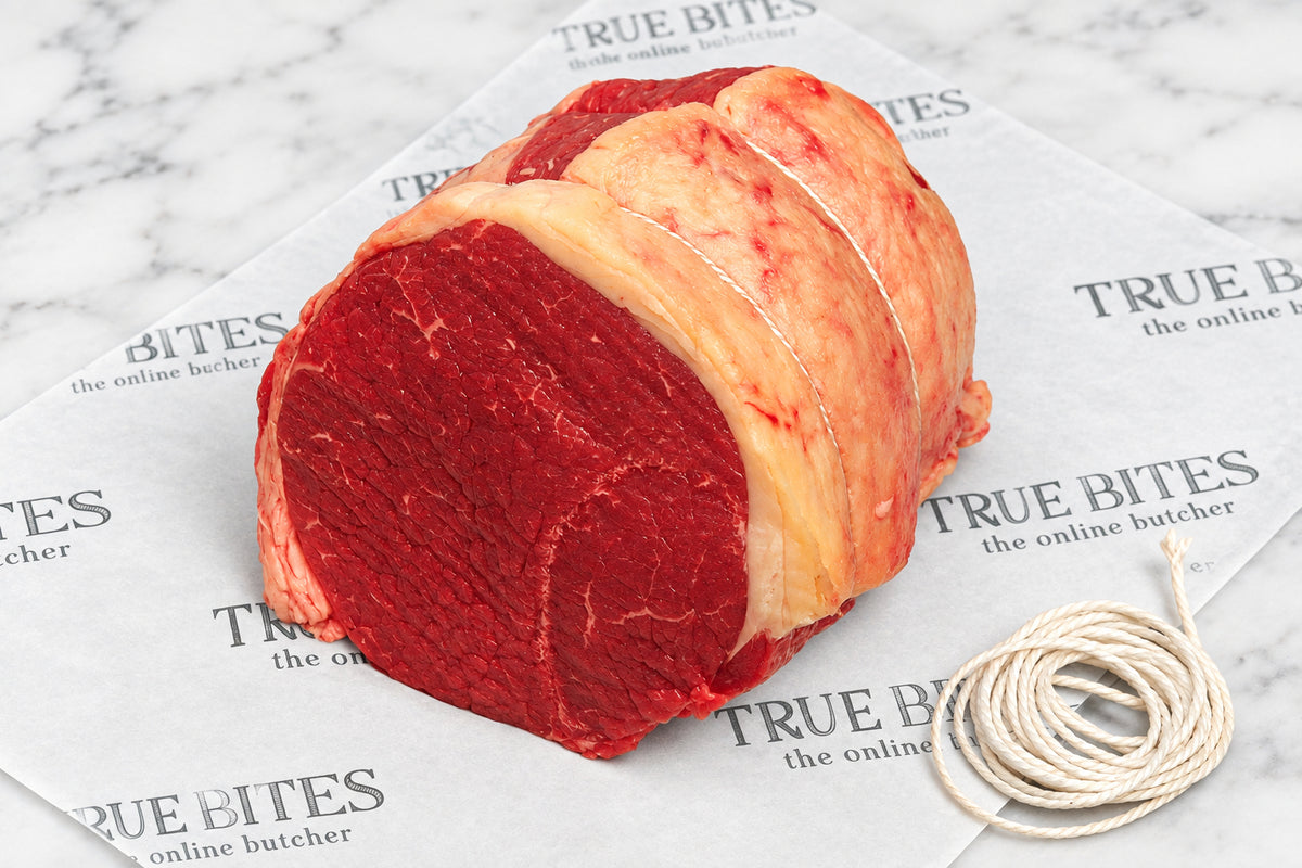 A raw beef silverside roast with fat wrapping, tied with string, placed on branded butcher paper over a dark wooden surface.