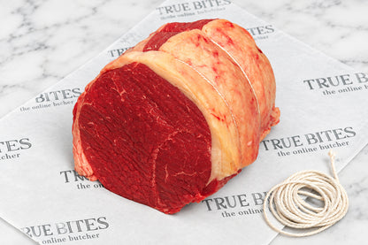 A raw beef silverside roast with fat wrapping, tied with string, placed on branded butcher paper over a dark wooden surface.