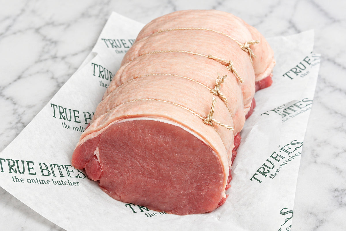 A raw, tied pork loin roast resting on branded butcher paper on a dark wooden surface, with a knife partially visible at the upper left.