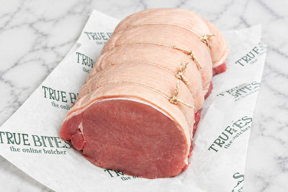 A raw, tied pork loin roast resting on branded butcher paper on a dark wooden surface, with a knife partially visible at the upper left.