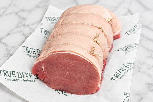 A raw, tied pork loin roast resting on branded butcher paper on a dark wooden surface, with a knife partially visible at the upper left.