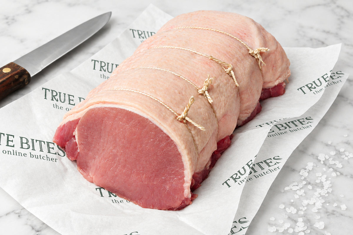 A tied raw boneless pork loin joint resting on branded butcher paper with a knife at the side on a dark wooden surface.