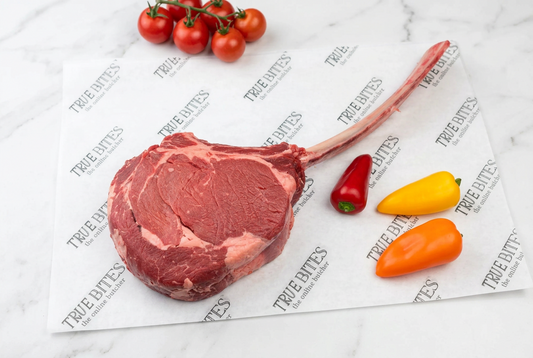 A raw tomahawk steak with a long bone, displayed on branded True Bites paper alongside three small peppers and a vine of cherry tomatoes on a white marble surface.