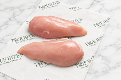 Two raw chicken breast halves placed on branded True Bites paper on a dark wooden table, with part of a knife visible at the top left.