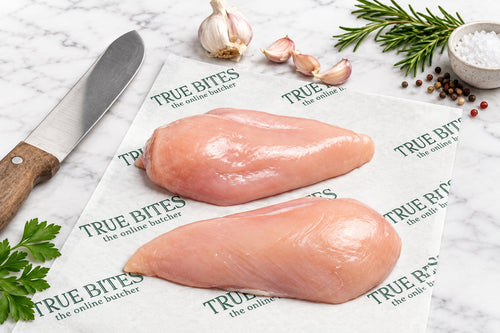 Two raw chicken breast fillets on branded paper next to a knife and garlic cloves on a dark wooden table.