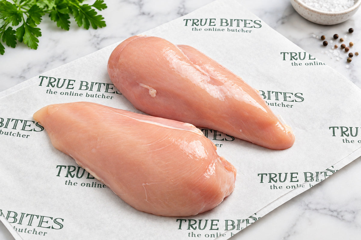 Two raw chicken breast fillets placed on branded True Bites butcher paper on a dark wooden surface, with a knife and garlic cloves visible around the edges.