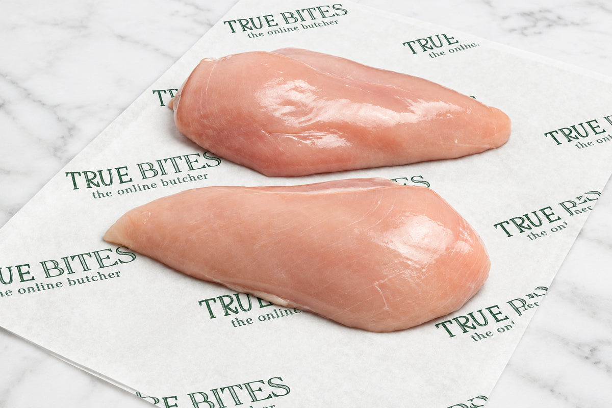 Two raw chicken breast fillets placed on branded paper wrapping, with garlic cloves and black pepper scattered nearby on a dark wooden surface. The paper shows the repeated brand text TRUE BITES the online butcher.