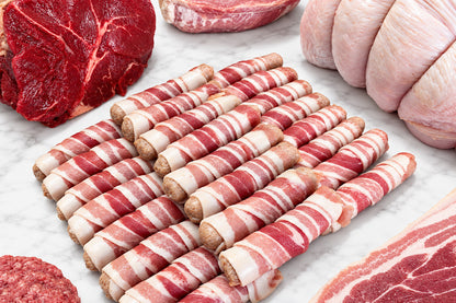 A close-up of 24 pigs in blankets arranged on a white surface, surrounded by raw meats including a red cut, a pale pink bundle, and marbled pork.