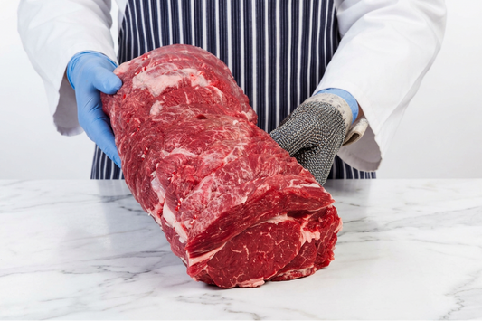 A large raw beef chuck roast with marbling is held upright by a person wearing a white coat, navy-and-white striped apron, blue gloves, and a steel chainmail glove on a white marble countertop.