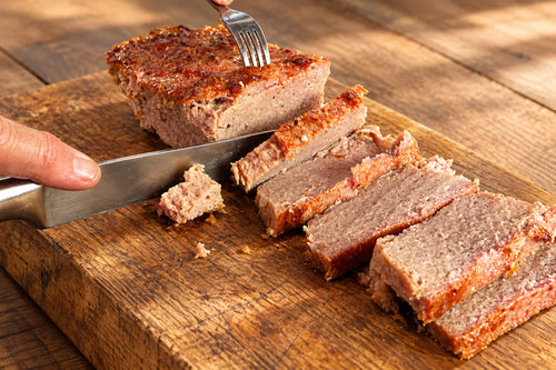 Cooked sausage meat slices on a wooden board, with a hand holding a knife and another hand using a fork in a kitchen setting.
