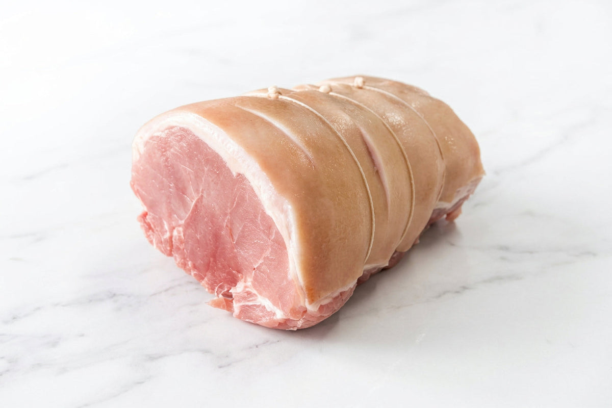 A raw, tied corner gammon roasting joint with rind on a white cutting board, with other raw meats blurred in the background.