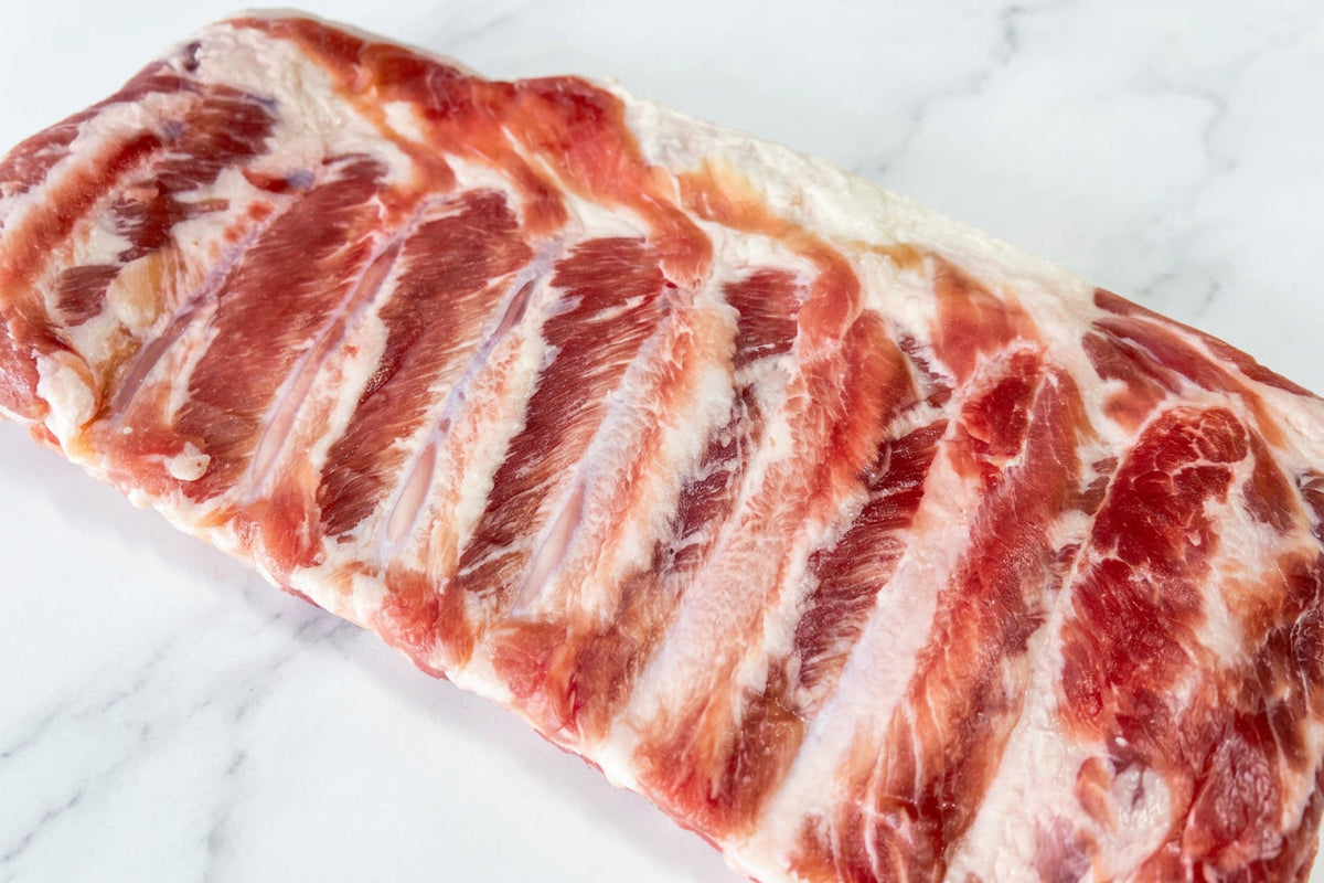 A close-up of a raw rack of cured pork bacon ribs with visible fat marbling on a plain white surface.