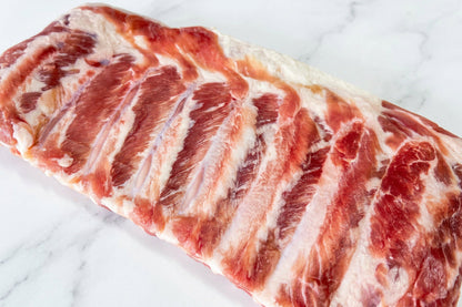 A close-up of a raw rack of cured pork bacon ribs with visible fat marbling on a plain white surface.