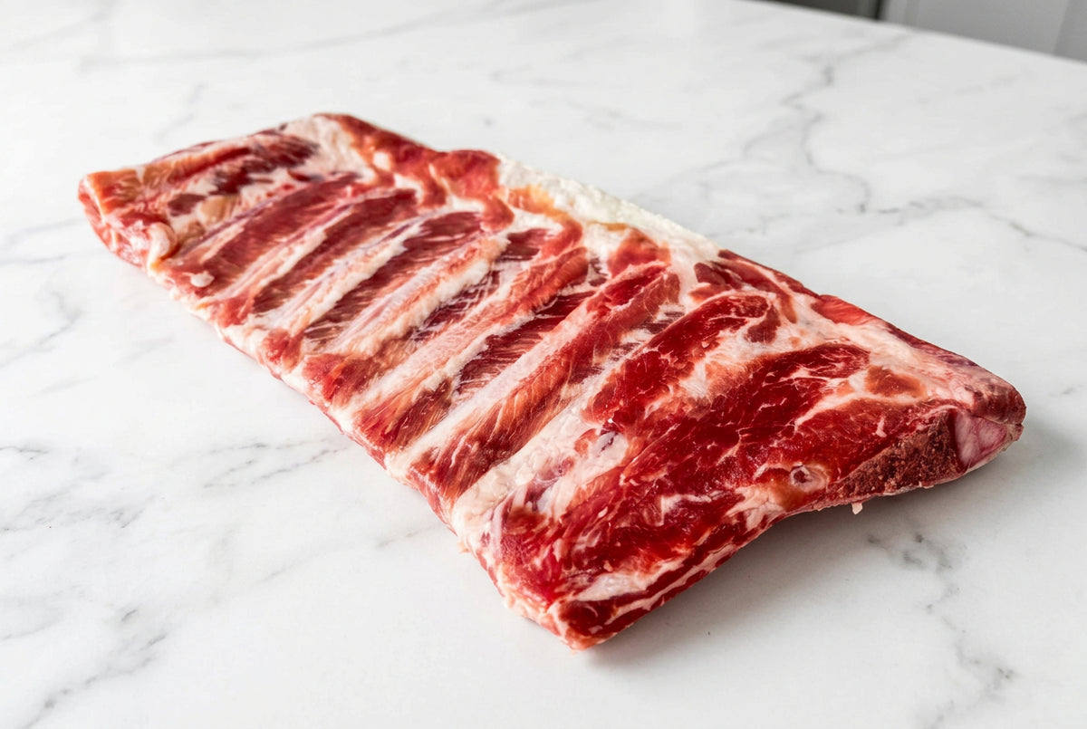 A raw rack of cured bacon ribs with visible marbling, placed on a clean white surface.