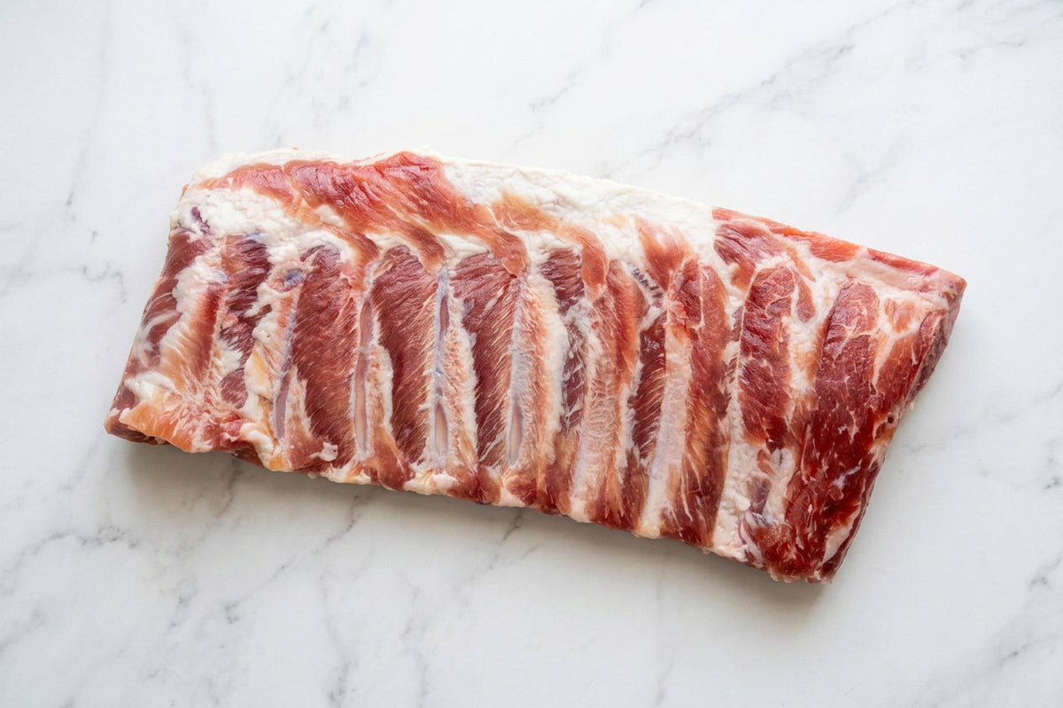 A raw rack of cured pork bacon ribs with visible marbling, photographed on a plain white surface.