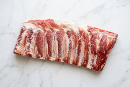 A raw rack of cured pork bacon ribs with visible marbling, photographed on a plain white surface.