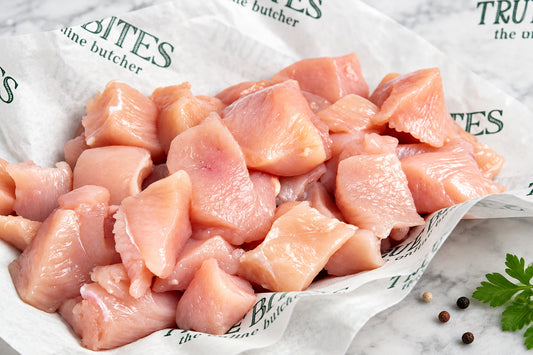 Raw, cubed chicken breast pieces piled on branded butcher paper over a dark wooden surface.