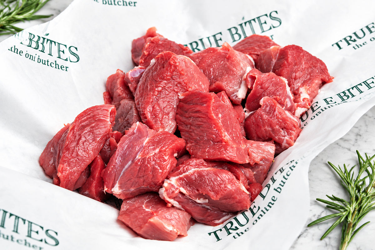 Raw diced lamb leg pieces piled on white butcher paper printed with the brand name 'True Bites the online butcher', with rosemary sprigs on a wooden surface around it.