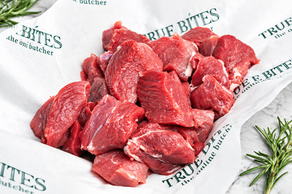 Raw diced lamb leg pieces piled on white butcher paper printed with the brand name 'True Bites the online butcher', with rosemary sprigs on a wooden surface around it.