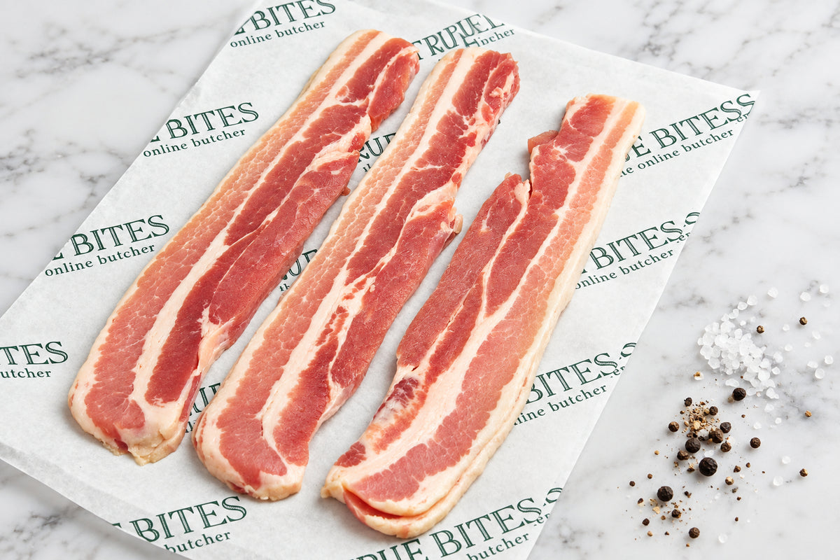 Strips of raw dry cured streaky bacon laid on branded paper reading 'TRUE BITES the online butcher' over a dark wooden surface, with coarse salt and peppercorns nearby.