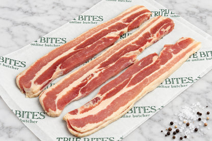 Strips of raw dry cured streaky bacon laid diagonally on branded paper reading 'TRUE BITES the online butcher' over a dark wooden surface, with coarse salt and pepper scattered nearby.