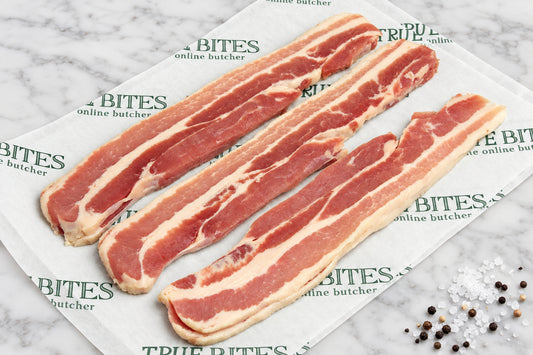 Strips of raw dry cured streaky bacon laid diagonally on branded paper reading 'TRUE BITES the online butcher' over a dark wooden surface, with coarse salt and pepper scattered nearby.
