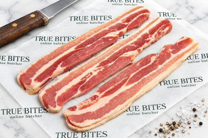 Strips of raw dry cured streaky bacon laid on branded paper reading 'TRUE BITES the online butcher' on a dark wooden surface, with a knife at the top and coarse salt at the bottom right.