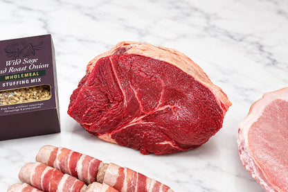 A raw beef rump roast with fat marbling, several pigs in blankets, a jar of cranberry sauce, and a boxed sage and onion stuffing on a white table in a kitchen or prep area.
