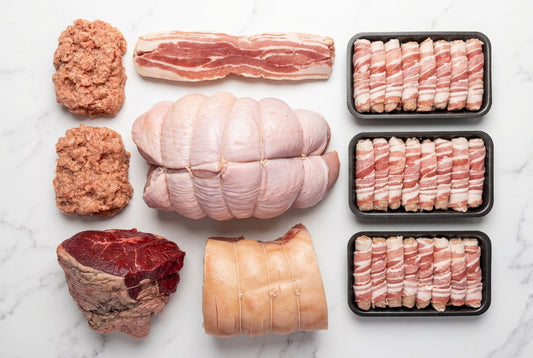 An assortment of Christmas meats arranged on a white background, including a beef rump roast, a gammon joint, pigs in blankets, streaky bacon, a tied whole turkey butterfly, two raw balls of sausage meat.