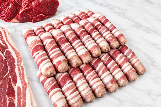 Raw pigs in blankets arranged on a white surface, with slices of marbled bacon and a cut of red meat visible at the edges.