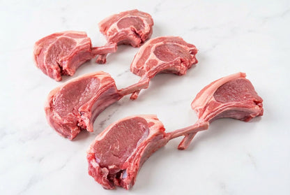 Six raw french trimmed lamb cutlets with bone, showing pink-red meat and white fat, arranged on a dark brown wooden table.