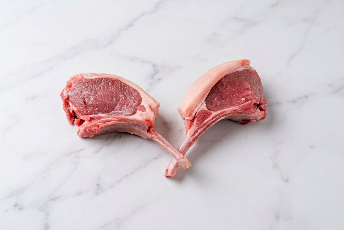 2 raw french trimmed lamb cutlets with bone, showing pink-red meat and white fat, arranged on a dark brown wooden table.