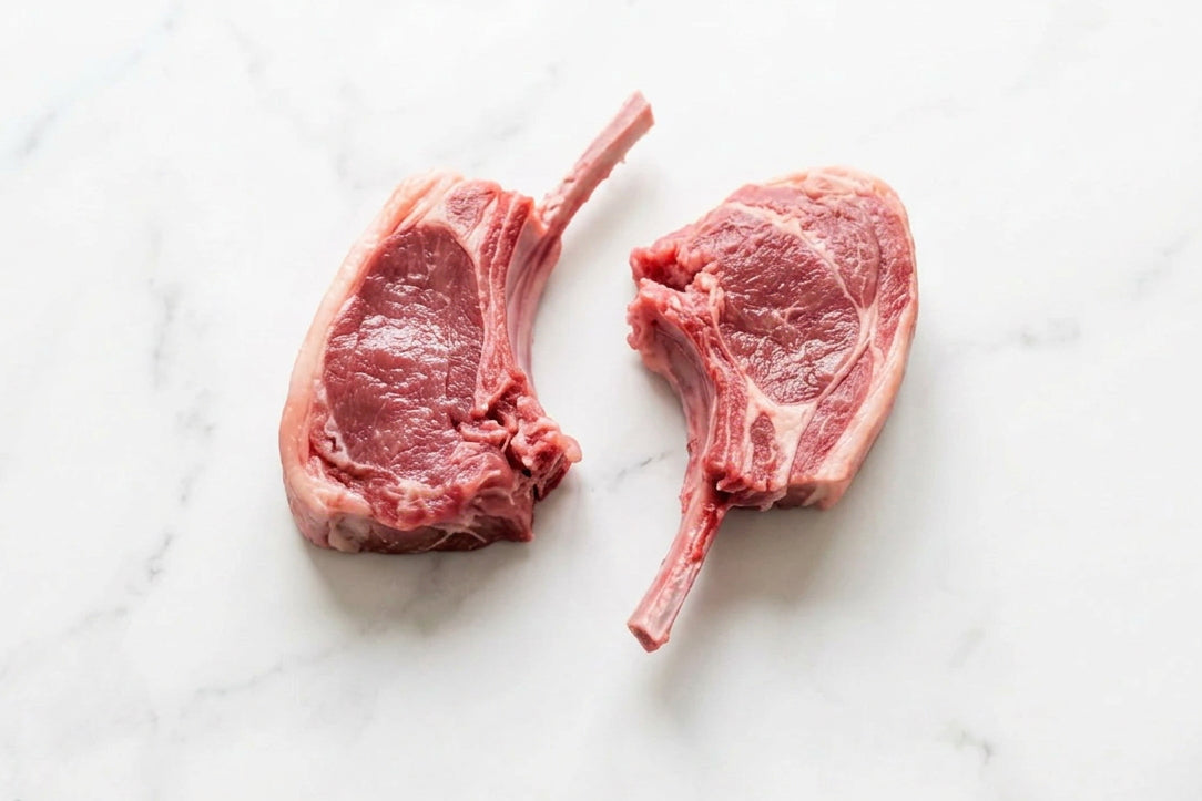 Four raw french trimmed lamb cutlets with bone, showing pink-red meat and white fat, arranged on a dark brown wooden table with 2 lamb cutlets cut off in the foreground.