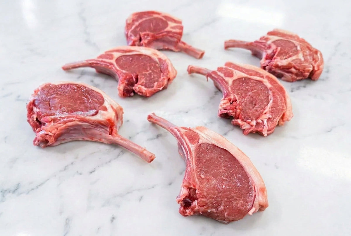Six raw french trimmed lamb cutlets with bone, showing pink-red meat and white fat, arranged on a dark brown wooden table.