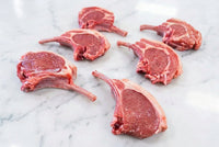Six raw french trimmed lamb cutlets with bone, showing pink-red meat and white fat, arranged on a dark brown wooden table.