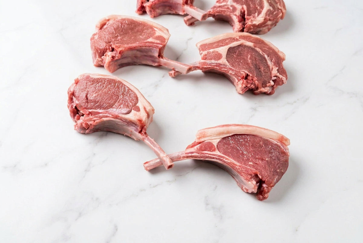 2 raw french trimmed lamb tomahawks in the centre with bone, showing pink-red meat and white fat, arranged on a dark brown wooden table.
