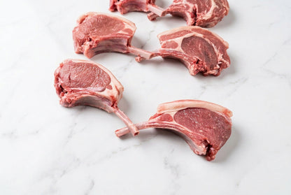 2 raw french trimmed lamb tomahawks in the centre with bone, showing pink-red meat and white fat, arranged on a dark brown wooden table.