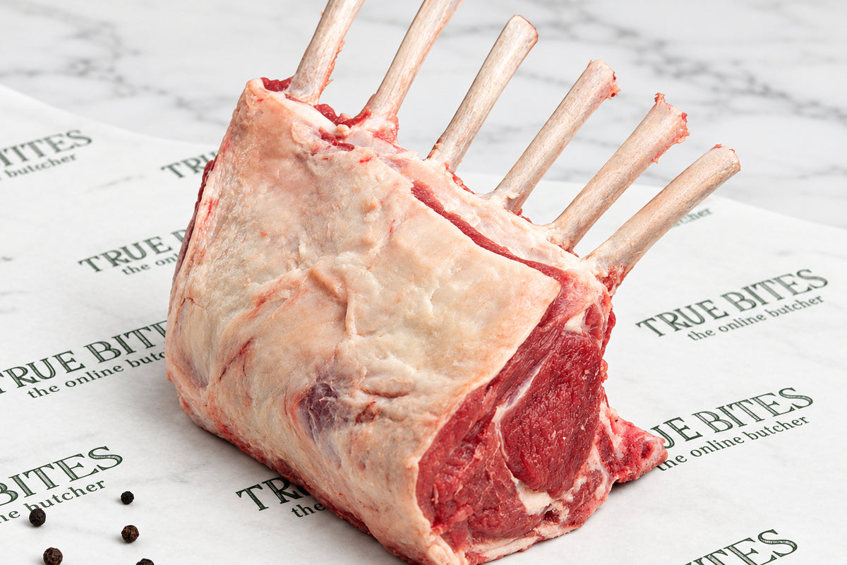 A raw French trimmed rack of lamb with bone, showing the marbled meat and fat, resting on branded butcher paper with peppercorns scattered nearby.