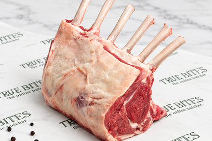 A raw French trimmed rack of lamb with bone, showing the marbled meat and fat, resting on branded butcher paper with peppercorns scattered nearby.