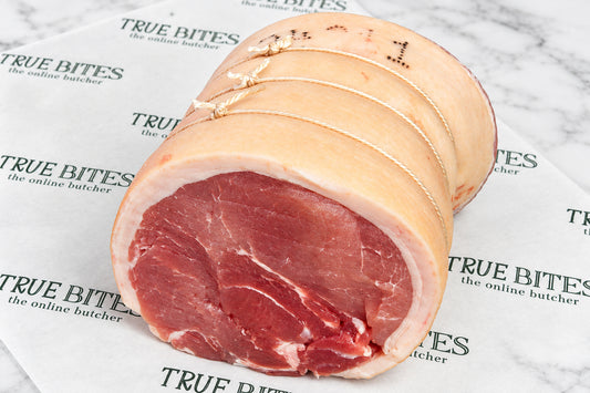 A rolled gammon horseshoe with white fat wrapping and tie marks, placed on branded butcher paper. The meat shows a deep red centre with white marbling, and the background includes a dark wooden surface and scattered salt.