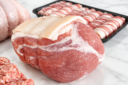 Raw gammon roasting joint on a white surface, including a large boneless turkey breast, a tray of pigs in blankets, and sausage meat in the foreground.