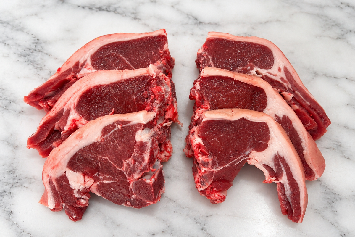 Four raw lamb chops with bone, showing deep red meat and white fat marbling, arranged on a white marble surface.