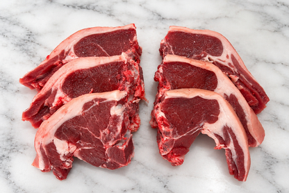 Four raw lamb chops with bone, showing deep red meat and white fat marbling, arranged on a white marble surface.