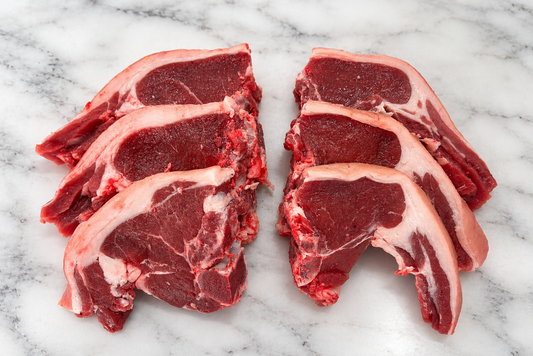 Four raw lamb chops with bone, showing deep red meat and white fat marbling, arranged on a white marble surface.