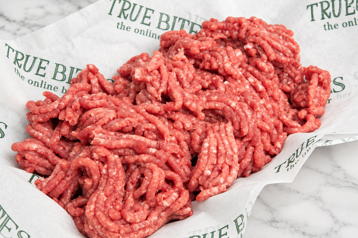 Raw ground lamb meat piled in a paper liner printed with the brand name True Bite, with a dark wooden surface and coarse salt visible in the corner.