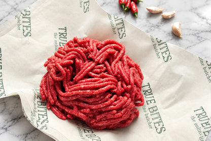 A mound of raw ground beef meat placed on branded paper wrapping on a wooden surface, with garlic cloves and red chili peppers nearby.